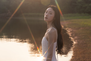 一位美丽的年轻女子穿着白色连衣裙站在靠近水域的地方 夕阳的光辉照亮了日落。