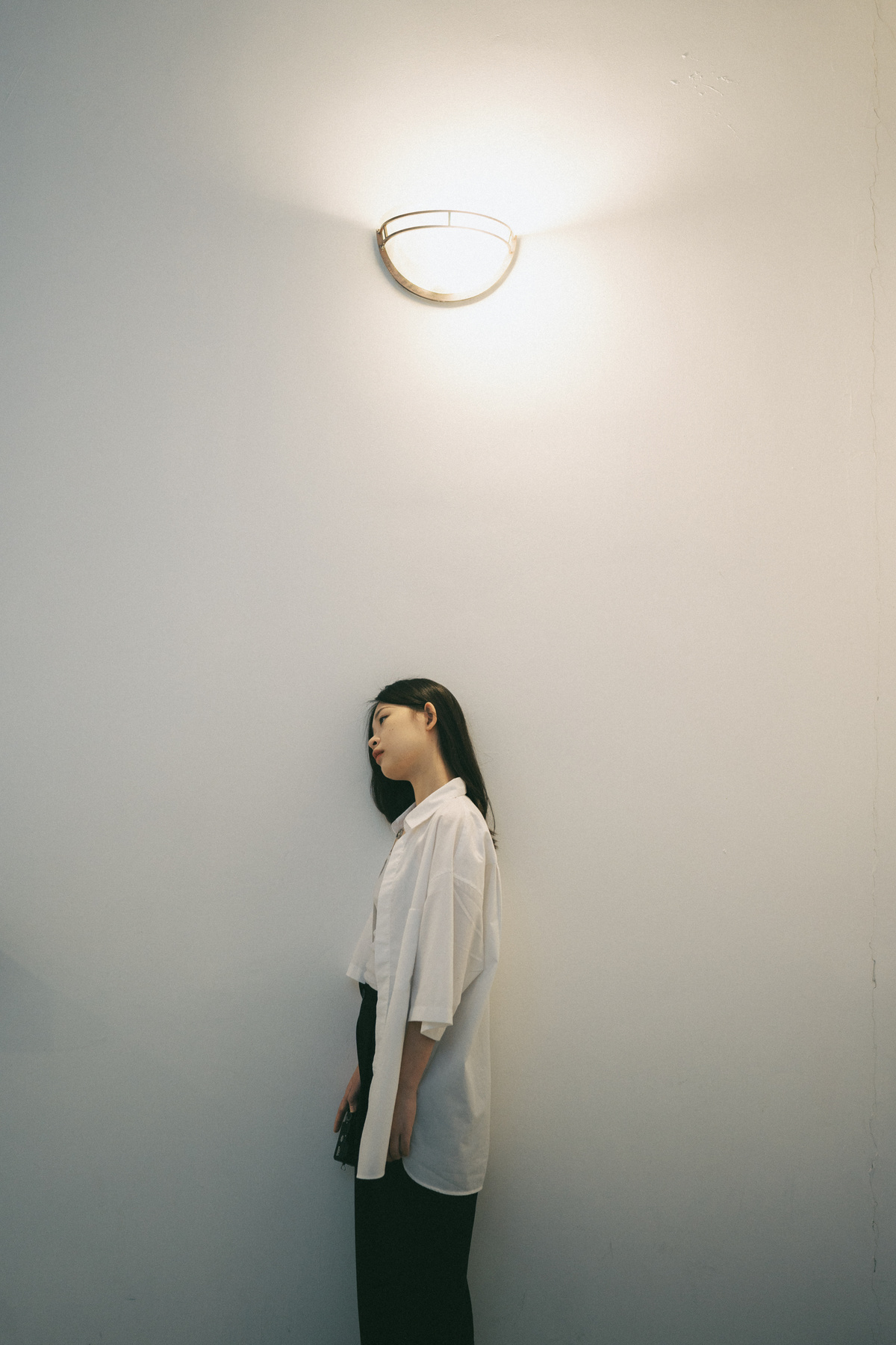 一位穿着白衬衫的年轻女子站在一面白墙上 她的头向一侧倾斜 墙上有一盏灯。