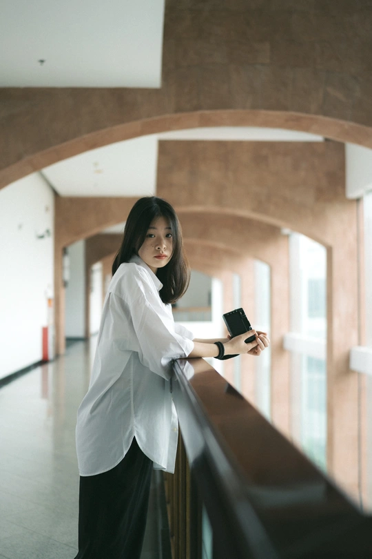 一位穿着白衬衫的年轻女子手握手机 倚靠在建筑物栏杆上