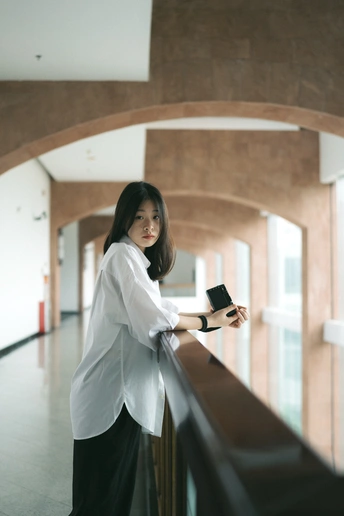 一位穿着白衬衫的年轻女子手握手机 倚靠在建筑物栏杆上