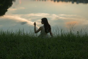一个年轻女子坐在湖边草地上 观赏日落时天空和水面的美景。