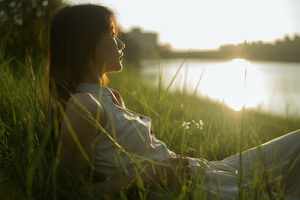 一位年轻女子在夕阳下坐在高草丛中 眺望着湖泊或河流。