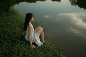 一位穿着白色连衣裙的年轻女子坐在湖边或河边草地上 仰望天空。