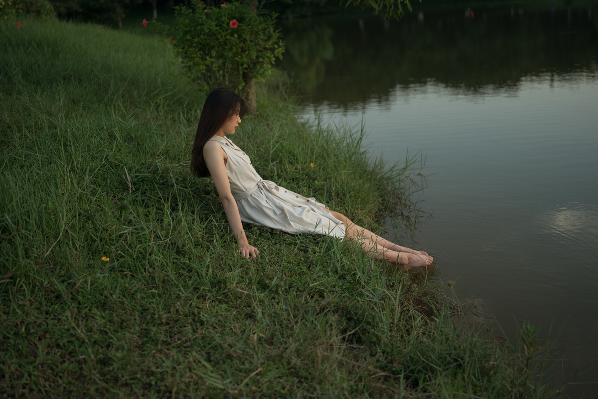 一个穿着白色连衣裙的年轻女子坐在水边或河边草地上