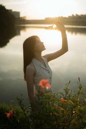 一位美丽的年轻女子手持一朵花 在夕阳西下时看着水面 太阳高高地挂在天空。