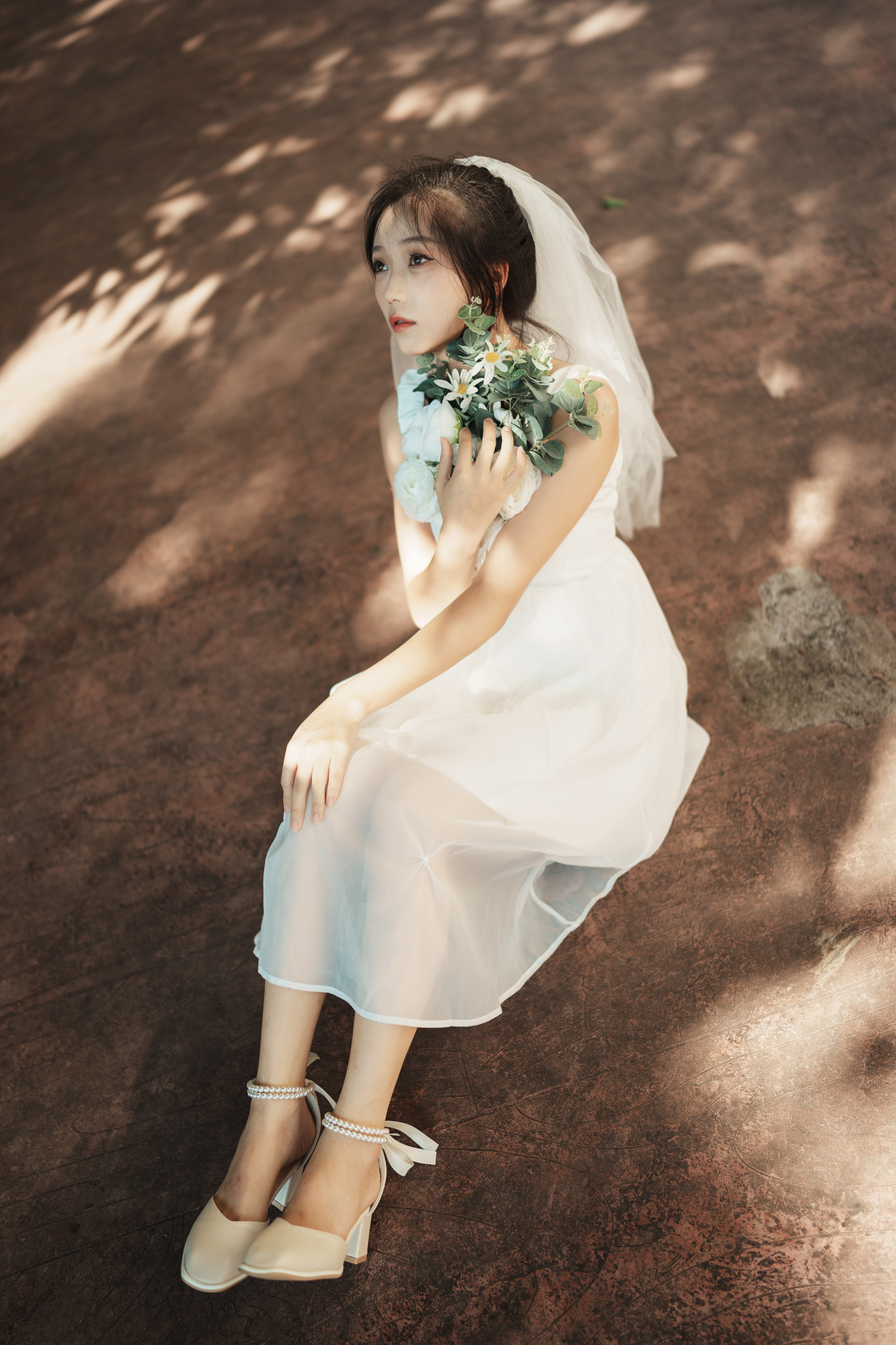 一位穿着白色连衣裙和白色鞋子的年轻女子抱着花坐在树下。