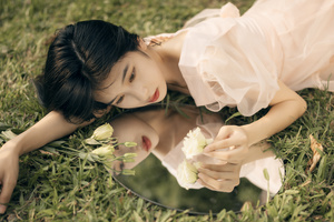 一个穿着白色裙子的年轻女子躺在草地上 看着一个有花放在上面的镜子。