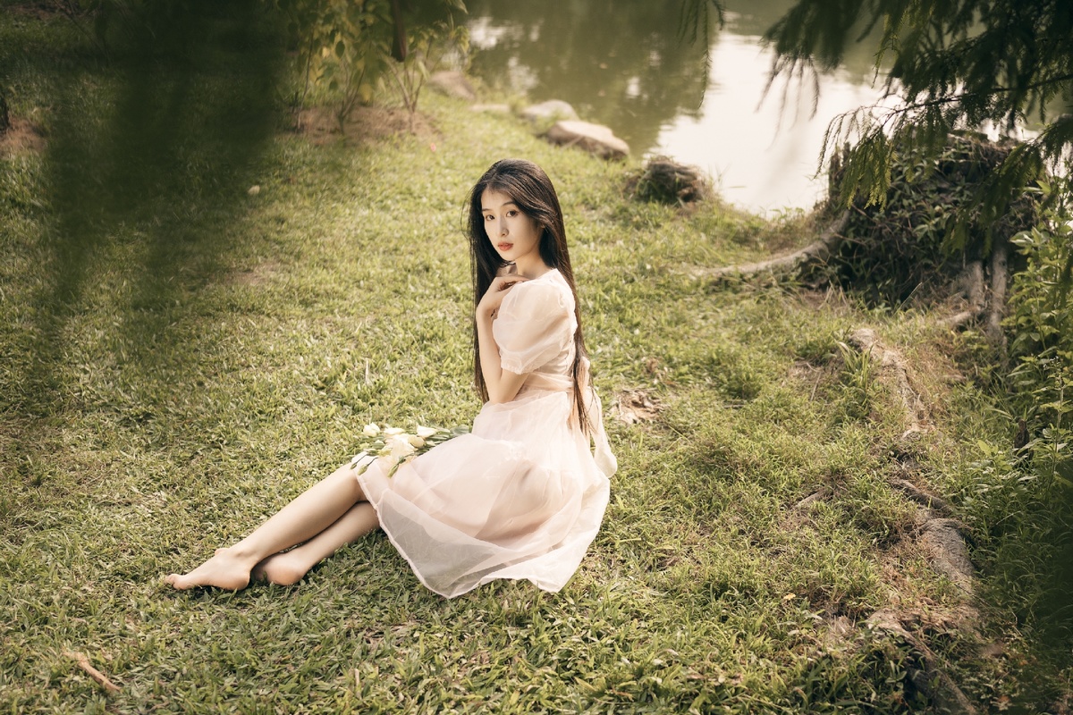 一位年轻美丽的女士穿着裙子坐在湖边或河边草地上 手里拿着一朵花。