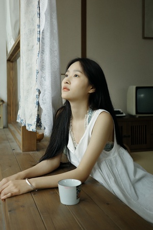 一个年轻女子坐在木地板上 正在透过窗户向外看 手里拿着一只白色杯子。