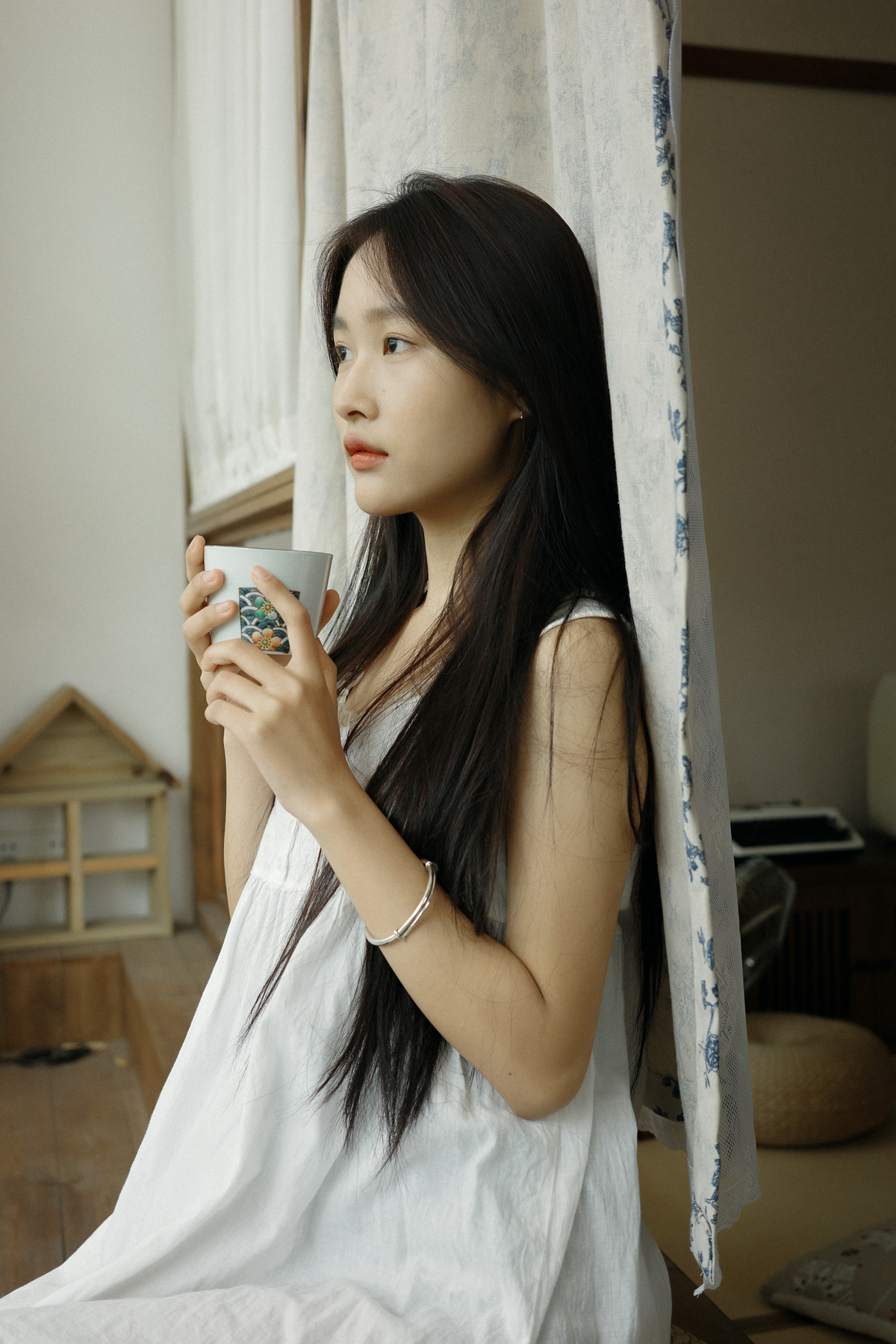 一位年轻女子拿着一杯茶或咖啡站在窗前 窗帘是透明的。