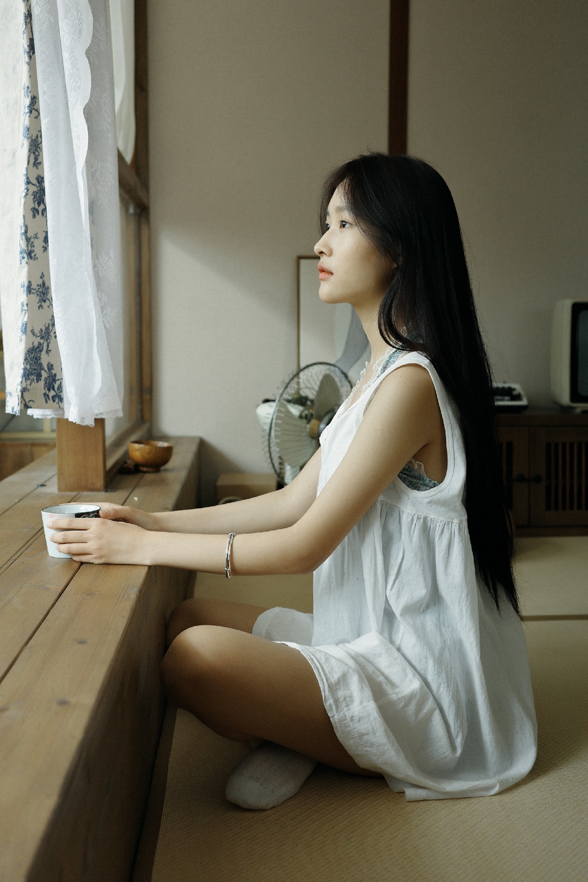一个穿着白色连衣裙的年轻女子坐在一个有窗户和茶杯的房间里的一张木制桌子上
