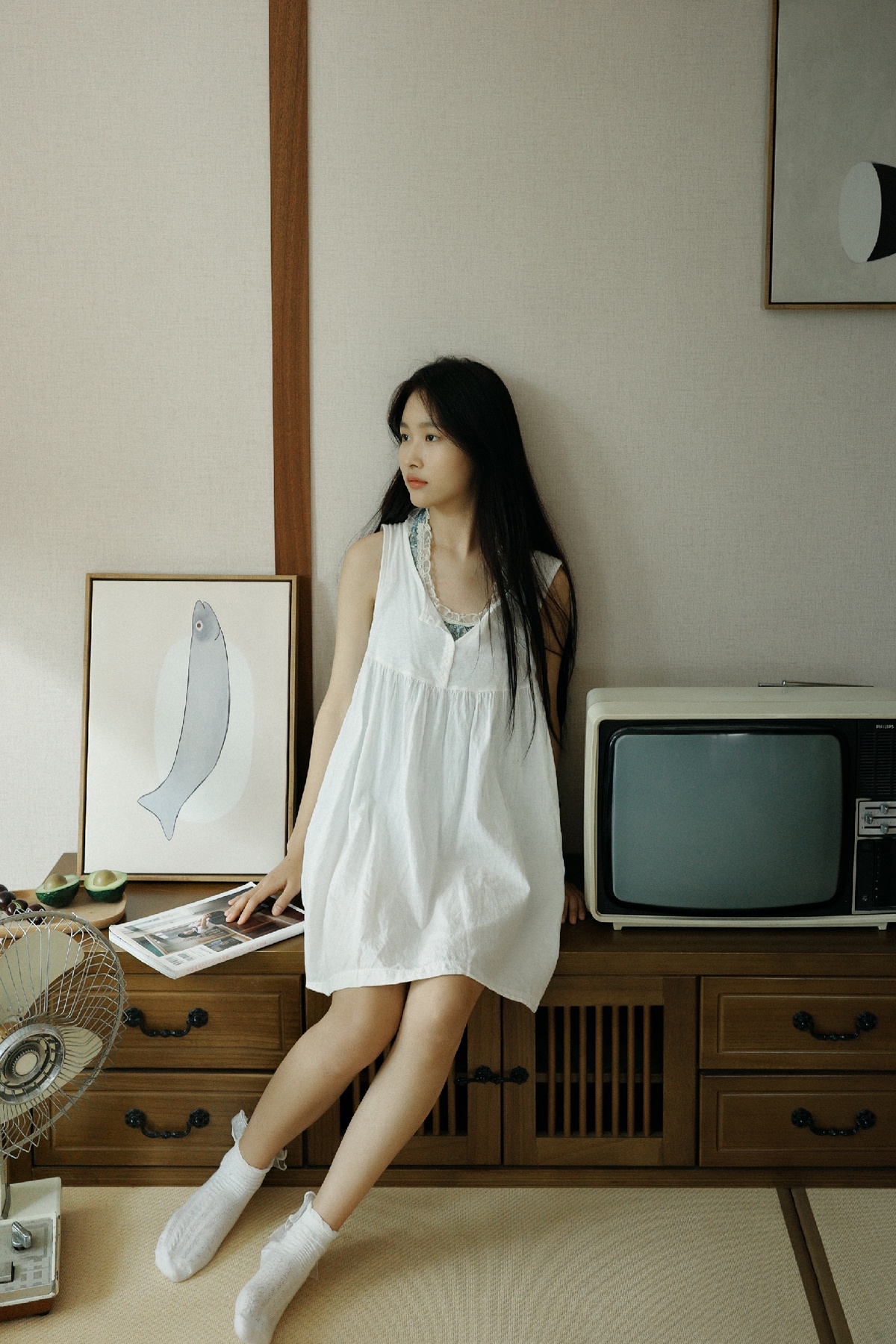 一个穿着白色连衣裙的年轻女子站在一个有电视和扇子的房间里