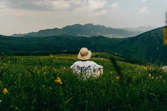 一个戴帽子和衬衫的年轻人坐在绿色的草地上 背景是山脉和丘陵 前景是一个人站着。