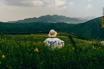 一个戴帽子和衬衫的年轻人坐在绿色的草地上 背景是山脉和丘陵 前景是一个人站着。