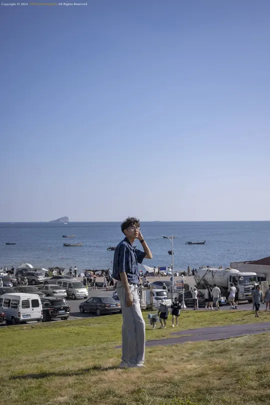 站在草地上 正在看手机的男子 前方是停满车的停车场 背景是海滩 海滩上有人们。