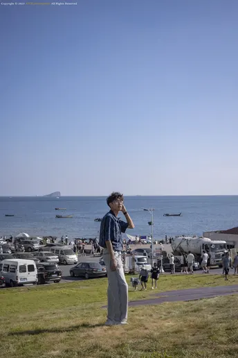 站在草地上 正在看手机的男子 前方是停满车的停车场 背景是海滩 海滩上有人们。