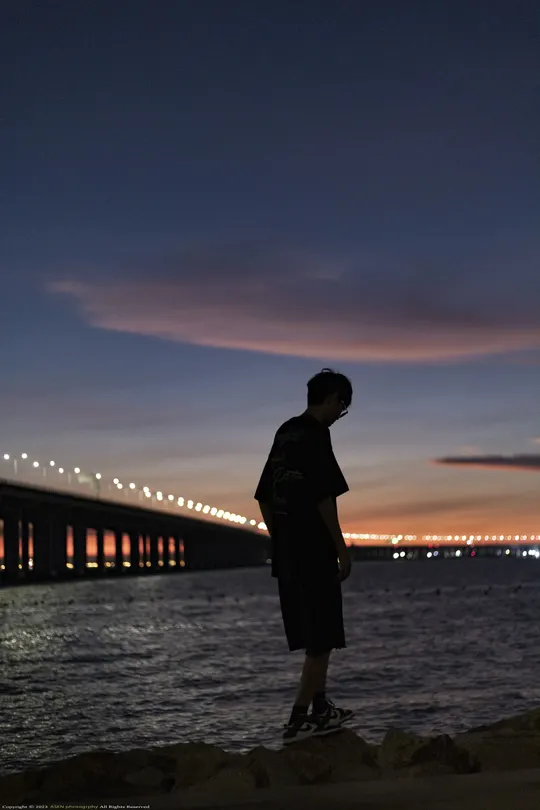 一个年轻男子站在海滩上 背景是夜晚的桥 正在注视着水面。