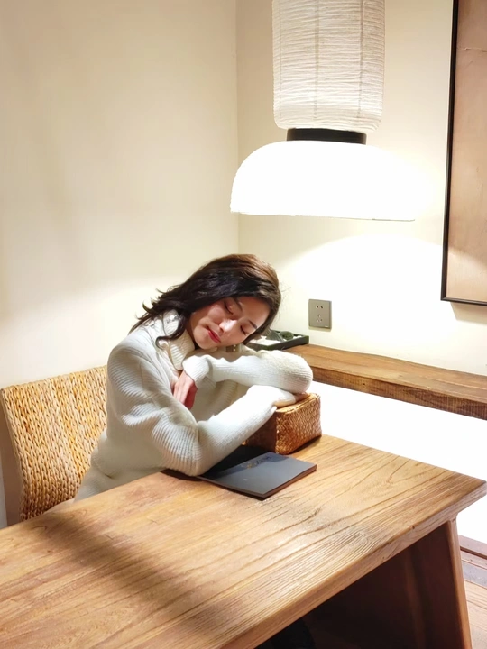 一个穿着白色毛衣的年轻女子坐在木制桌子旁 旁边有一台笔记本电脑和一台平板电脑。