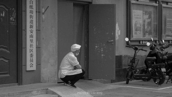 一张黑白照片 一位老妇人坐在人行道上 前方有一栋建筑 街道上停着摩托车。