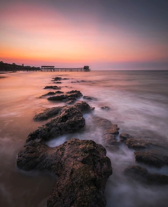 夕阳或日出时 远处有码头 沙滩上有岩石 太阳落下的天空
