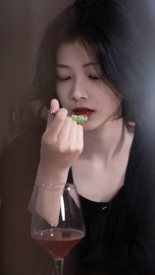 一个涂着红唇的女人正在桌子旁吃西兰花 并喝着一杯酒。