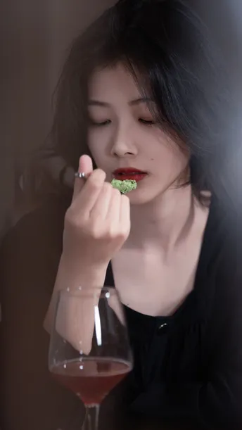 一个涂着红唇的女人正在桌子旁吃西兰花 并喝着一杯酒。
