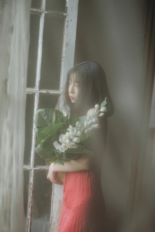 一位穿着红裙子的年轻女子抱着一束白色花 正在透过窗户向外看。