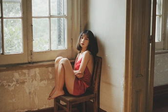 一个穿着红裙子的年轻女子坐在木椅上 靠在窗户边。