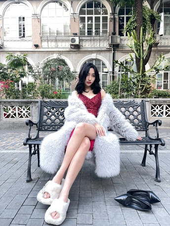 一位穿着红色连衣裙和白色皮草大衣的女人坐在长凳上 双腿交叉。