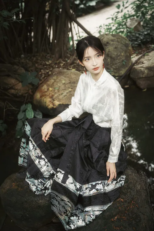 一个穿着白色连衣裙和黑色裙子的年轻女子坐在河中的一块岩石上