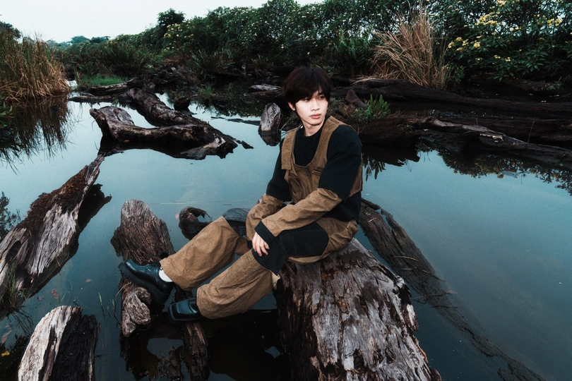 一个年轻人坐在一条小溪的树干上 背景中另一个人坐在岩石上。