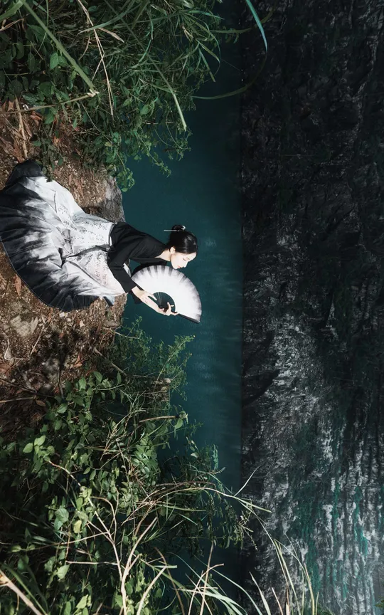 一位穿着黑白裙子的女士站在水边 手里拿着一把雨伞。