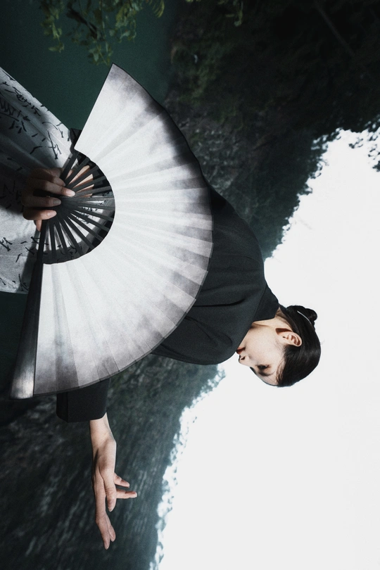 一位穿着黑白裙子的年轻女子手持扇子站在水边