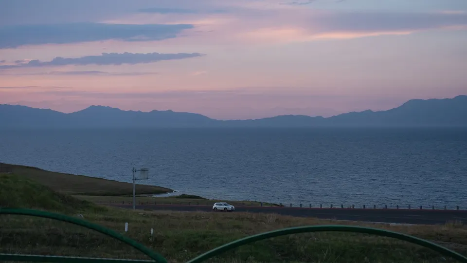 日落时分的湖泊 远处有山脉 道路后方有栅栏和一辆汽车。