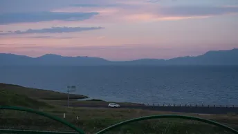 日落时分的湖泊 远处有山脉 道路后方有栅栏和一辆汽车。