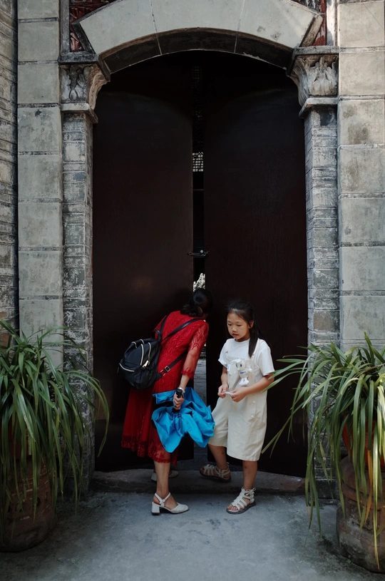 一个背着书包的女孩站在一栋建筑物的门口 而她的母亲走了过去。