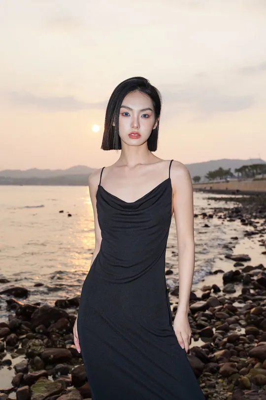 一个穿着黑色连衣裙的年轻女子站在日落时分的岩石海滩上