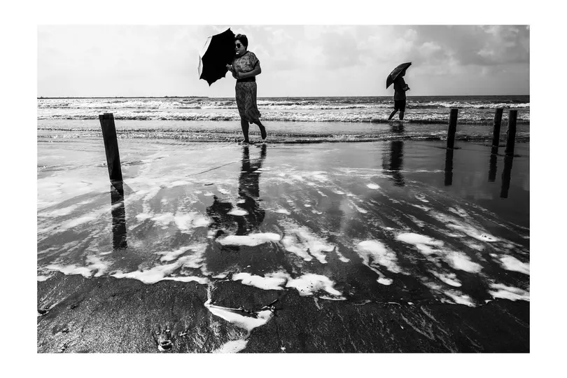 一个穿着黑白色衣服的男子打着伞在沙滩上行走 而人们站在水中。