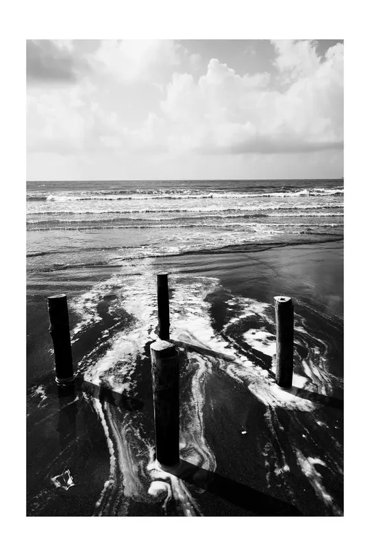 一张黑白照片 前景是沙滩、沙子和杆子 背景是水和波浪。