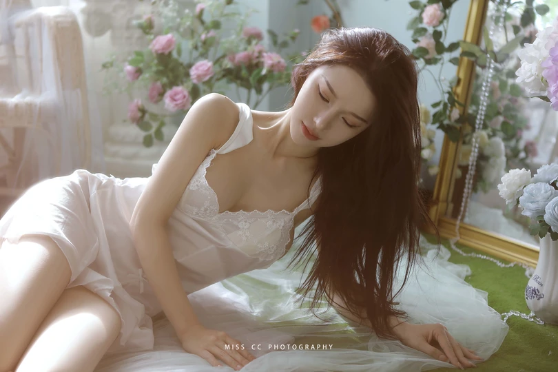 一个穿着白色连衣裙的年轻女子躺在床上 床前有花 镜子里反射着她的身影。