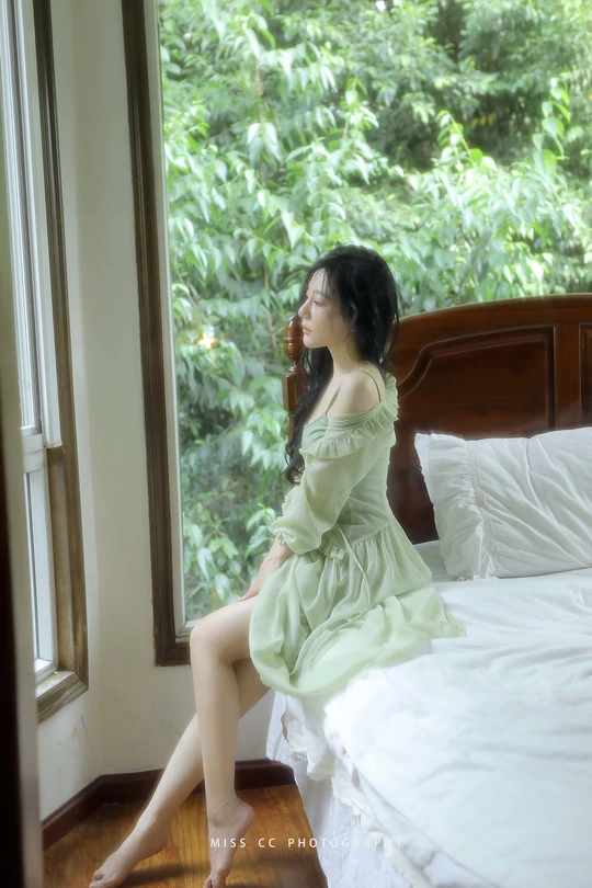 一个穿着绿色连衣裙的年轻女子坐在床上 凝视着窗户。