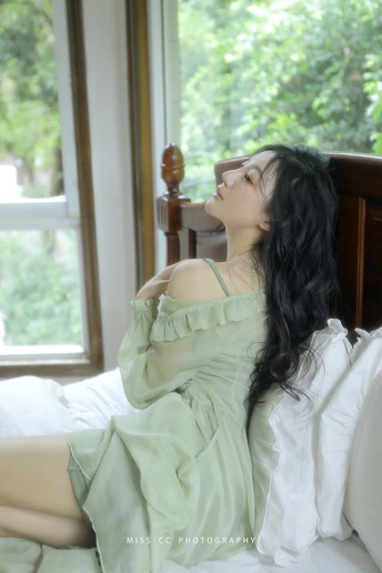 一个穿着绿色连衣裙的年轻女子坐在床上 凝视着窗户。