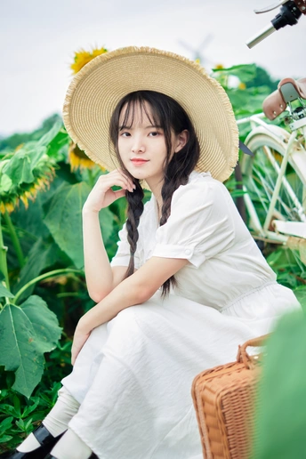 一位身穿白色连衣裙、头戴草帽的年轻女子坐在向日葵地里的自行车上