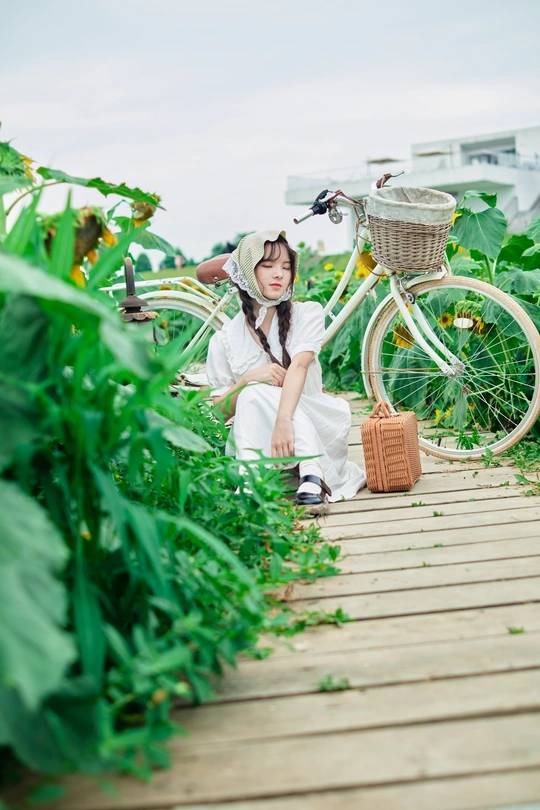 一个穿着白色衣服的年轻女孩坐在一座木桥上 旁边是一辆提着篮子的自行车