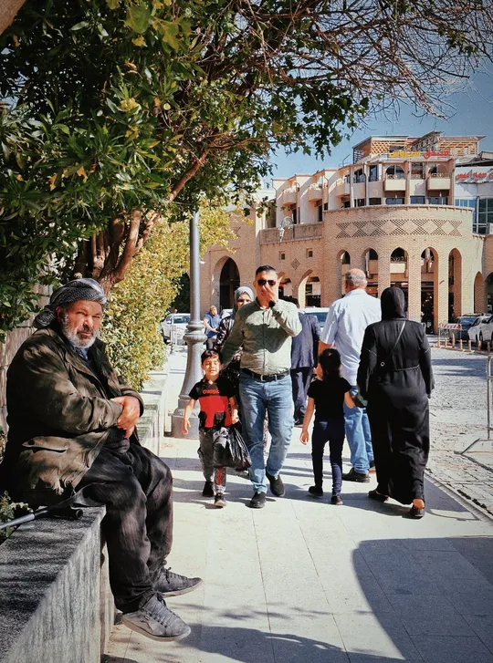 一位老人坐在城市人行道上的长椅上 周围有树和其他人。