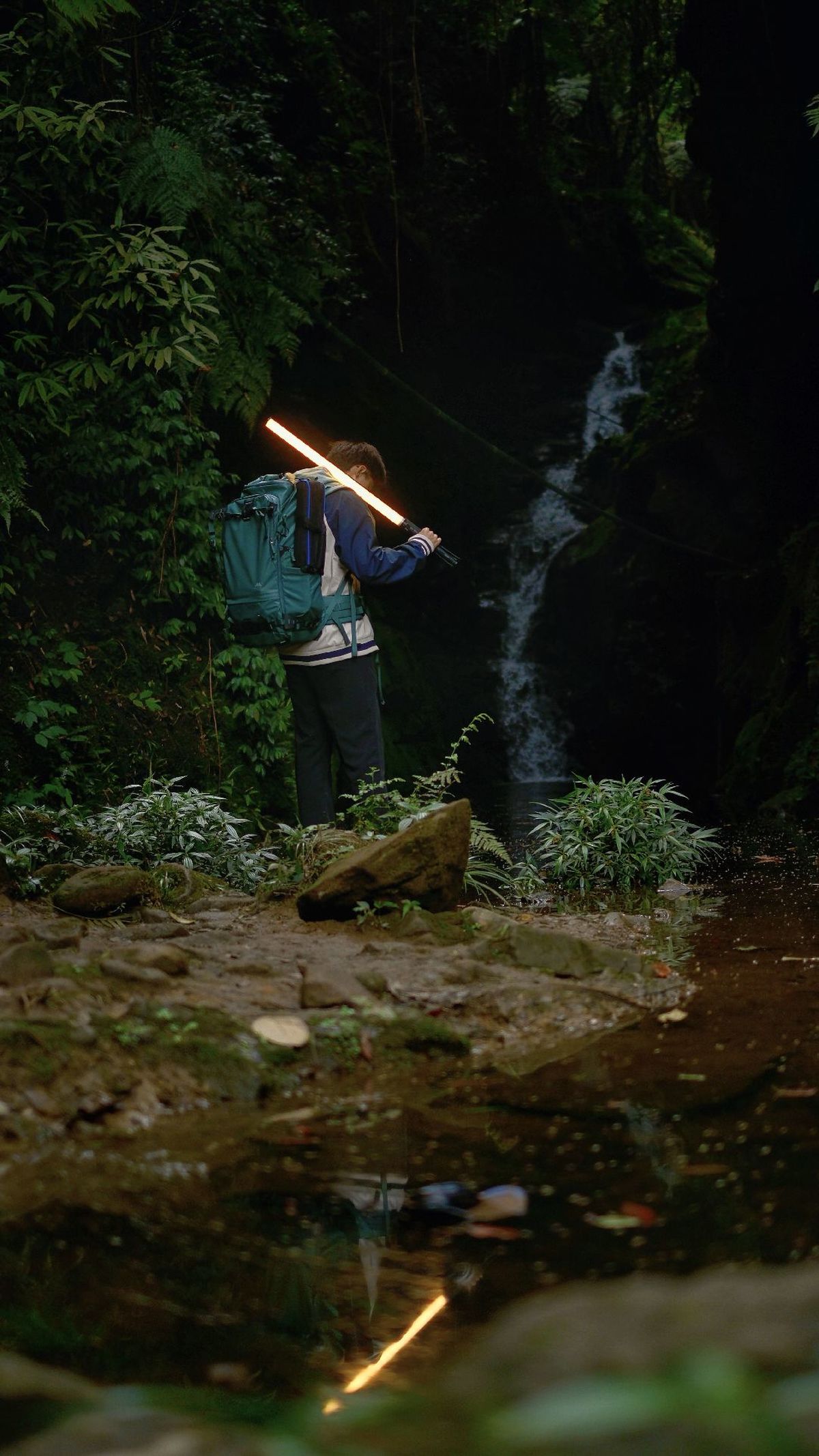 一个背着书包的男子站在夜晚的森林小溪旁 手里拿着手电筒。