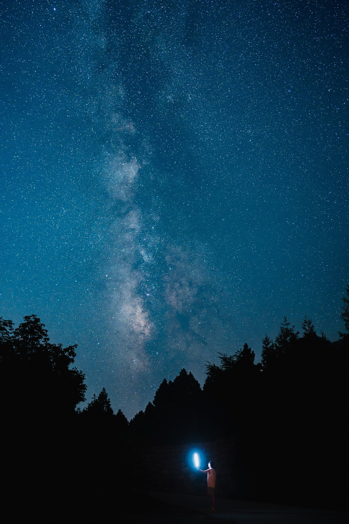 一个人站在森林里夜晚 手持手电筒照向地面 黑暗的蓝色天空布满星星。