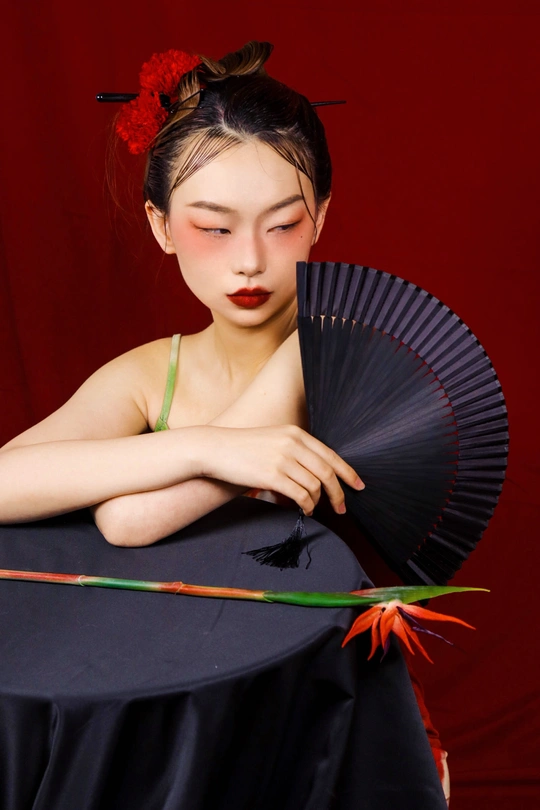 一位年轻女子手持红花扇子坐在黑色桌子旁