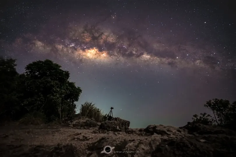 一个人站在夜晚的自行车旁边 天空中繁星闪烁 树木环绕。