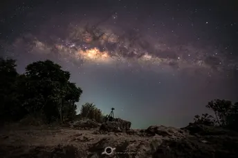 一个人站在夜晚的自行车旁边 天空中繁星闪烁 树木环绕。